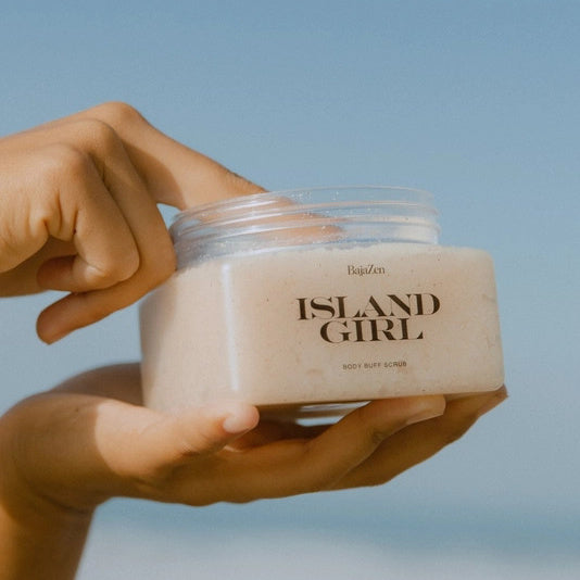 Hand holding a jar of 'Baja Zen Island Girl' body scrub against a blue sky background