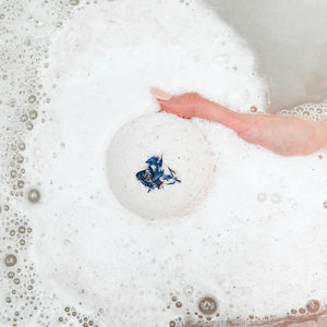 Bath bomb with purple flower inside being dropped into a bathtub filled with bubbles.