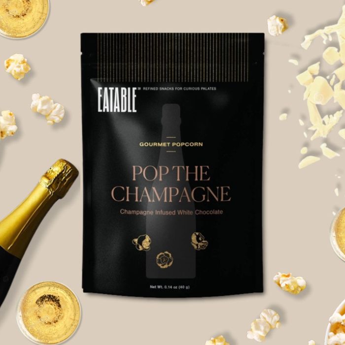 Eatable gourmet popcorn bag in Pop The Champagne flavor surrounded by popcorn and a bottle of champagne on a light background