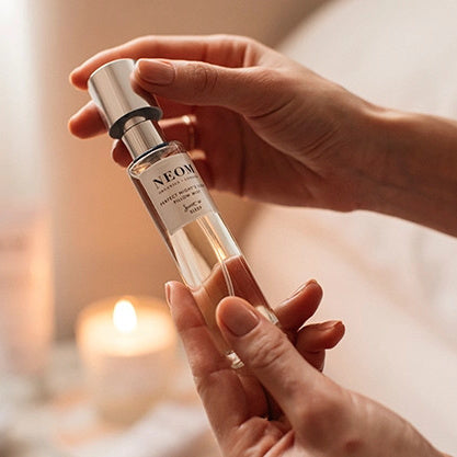 Person holding a Neom Wellbeing Perfect Night's Sleep Pillow Mist bottle with a blurred candle in the background