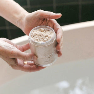 Person holding a jar of Palermo Body Soothing Milk Bath powder over a bathtub with a blurred background