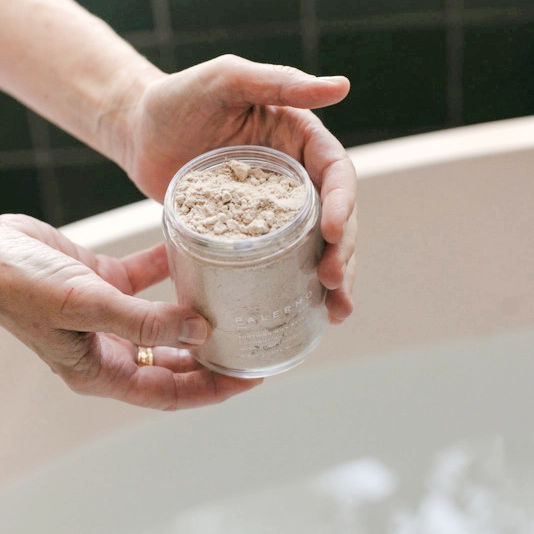 Person holding a jar of Palermo Body Soothing Milk Bath powder over a bathtub with a blurred background