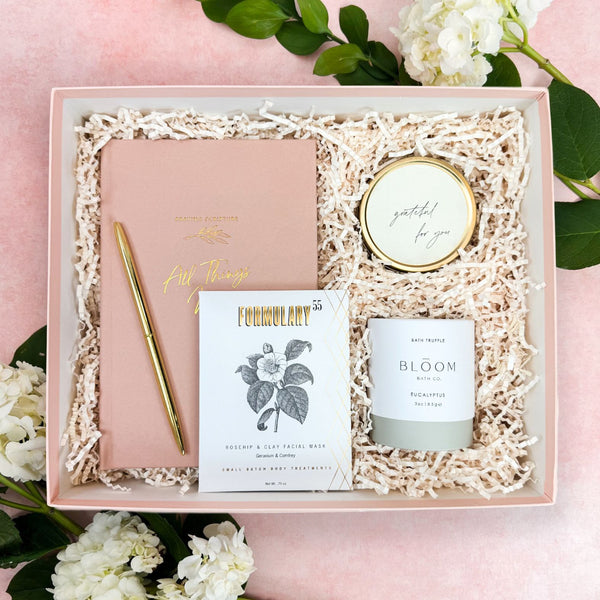 A Christian curated gift box set arranged in a blush box filled with ivory shredded paper. The set includes a blush pink Praying Scripture journal with gold lettering, a gold pen, a white and gold candle that says 'grateful for you', a Bloom Bath Co. Eaucalyptus Bath Truffle, and a Formulary 55 Rosehip & Clay Facial Mask.