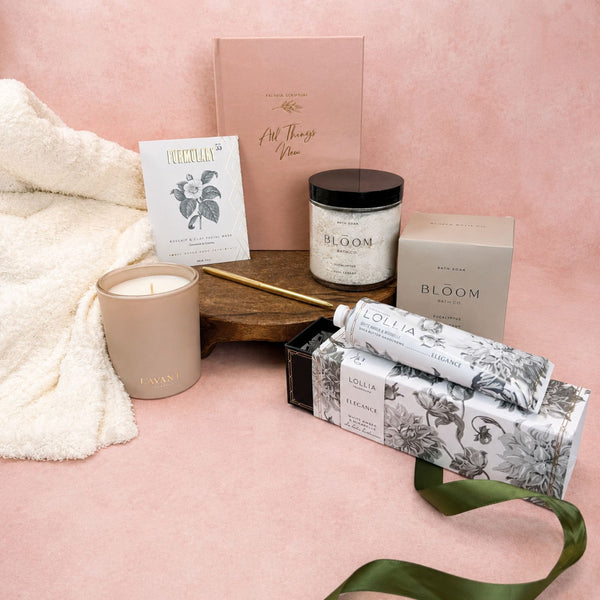 Deluxe All Things New Christian gift box arranged with a buttery soft ivory throw blanket, a Praying Scripture journal, a gold pen, a Formulary 55 Rosehip & Clay Mask, a Lollia Elegance Shea Butter Handcreme with a green ribbon, a L'AVANT blushed bergamot candle, and a Bloom Bath Co. Eucalyptus bath soak, set against a pink background.