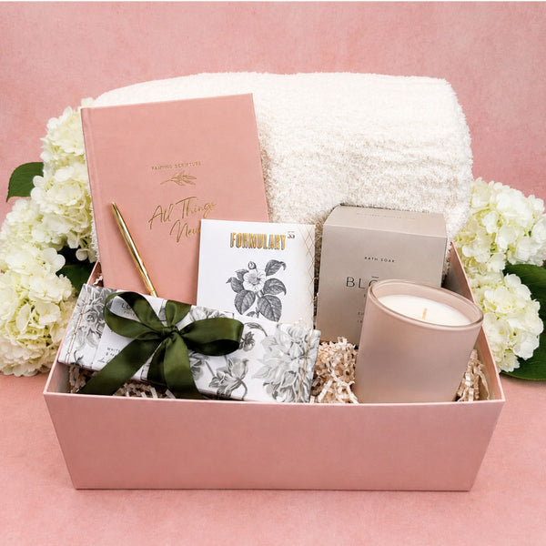 Deluxe All Things New Christian gift box arranged with a buttery soft ivory throw blanket, a Praying Scripture journal, a gold pen, a Formulary 55 Rosehip & Clay Mask, a Lollia Elegance Shea Butter Handcreme with a green ribbon, a L'AVANT blushed bergamot candle, and a Bloom Bath Co. Eucalyptus bath soak, set against a pink background with white hydrangea flowers.
