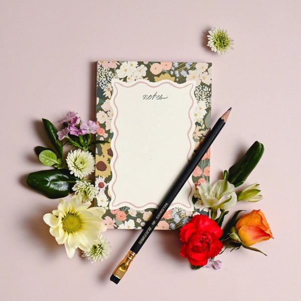 Amy Heitman Flower Market notepad with a cream center writing area framed by a multicolor floral border, styled on a flatlay with a black pencil and small flowers around it on a solid pale lavender background | Build A Luxury Custom Gift Box for Women with Luxe & Bloom