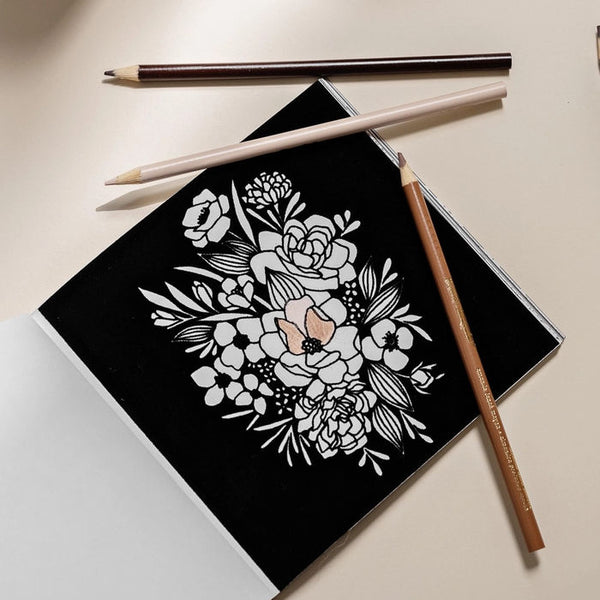 Bloom coloring book with floral design and three colored pencils on a light surface