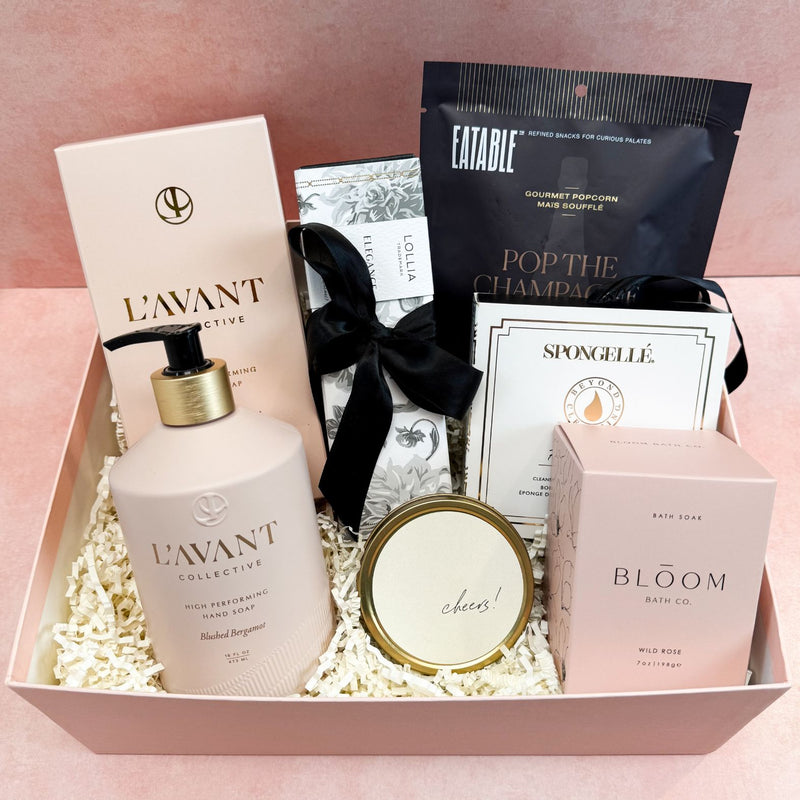 Blush & Bubbly Luxury Curated Gift Box with self care items such as L'avant Collective Blushed Bergamot Hand Soap, Lollia Ellegance Shea Butter Hand Cream, Bloom Bath Co. Wild Rose Bath Soak, Spongellé Freesia Pear Boxed Body Buffer, Cheers Tin Candle, and Pop The Champagne Popcorn | Luxury Curated Gift Boxes for Women from Luxe & Bloom