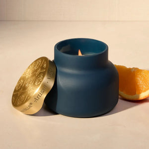 A matte navy blue Capri Blue Volcano Terrazza Velluto Petite Candle jar with a small flame, shown with its gold lid leaning beside it on a light surface. A slice of orange is placed behind the candle.