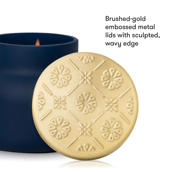 A round brushed-gold metal candle lid with embossed floral and geometric patterns, shown in front of a dark navy blue candle container. Text on the right reads, 'Brushed-gold embossed metal lids with sculpted, wavy edge.