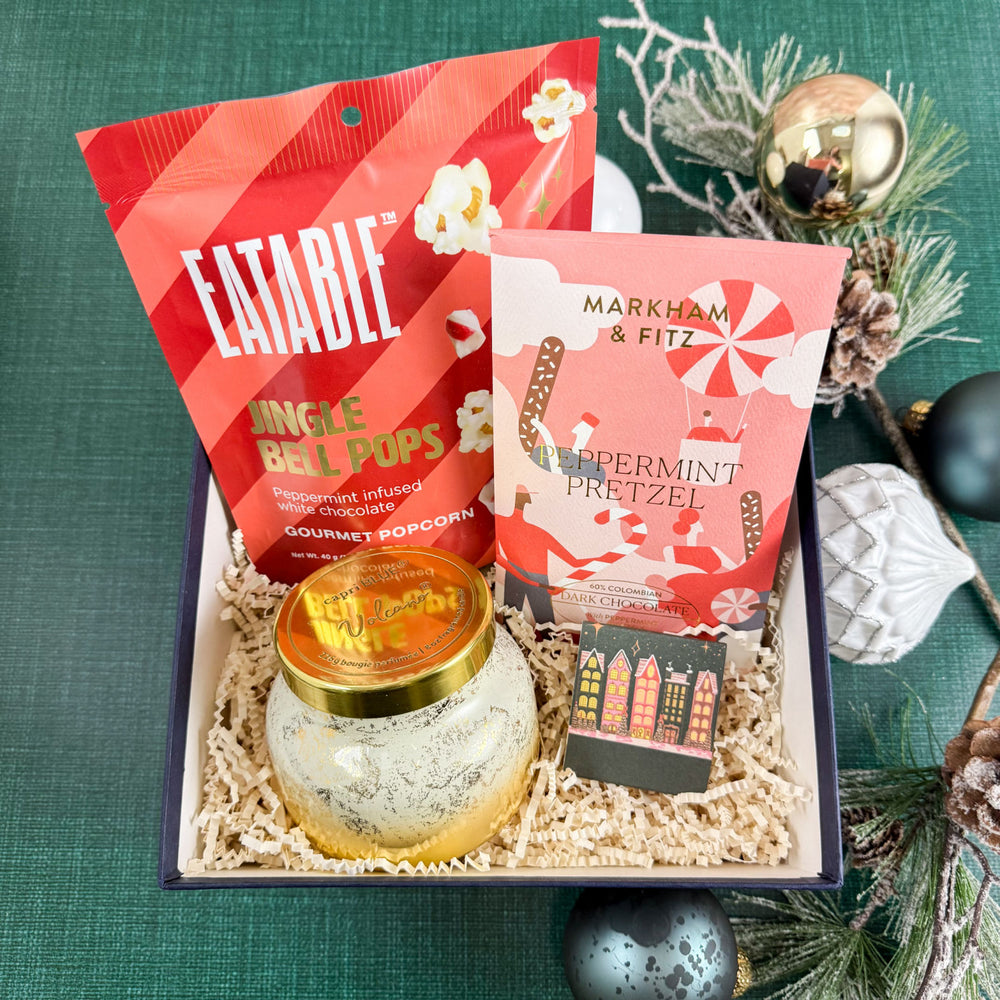 Comfort & Joy Luxury Curated Christmas Gift Box with Eatable Jingle Bell Pops Popcorn, Markham & Fitz Peppermint Pretzel Chocolate Bar, Capri Blue Volcano Glimmer Candle, and Wintry Village Printed Matchbook on a green textured background