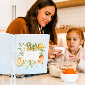A woman and a young girl are baking in a bright kitchen, with the open cover of a family recipe book displayed in the foreground. The book has a light blue cover decorated with watercolor illustrations of citrus fruits and green leaves, and the title 'family recipe book' is visible.