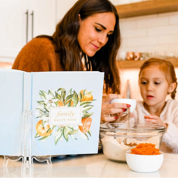 A woman and a young girl are baking in a bright kitchen, with the open cover of a family recipe book displayed in the foreground. The book has a light blue cover decorated with watercolor illustrations of citrus fruits and green leaves, and the title 'family recipe book' is visible.