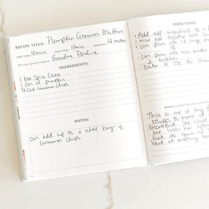 An open recipe notebook showing two pages with handwritten notes for pumpkin cinnamon muffins, photographed on a light marble surface.