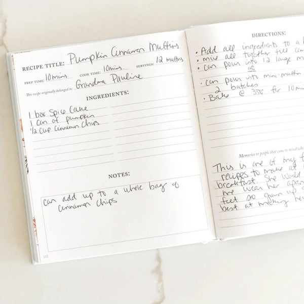 An open recipe notebook showing two pages with handwritten notes for pumpkin cinnamon muffins, photographed on a light marble surface.