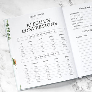 An open recipe book showing printed kitchen conversion cheat sheets, including liquid and dry measurement tables, photographed on a light marble surface.