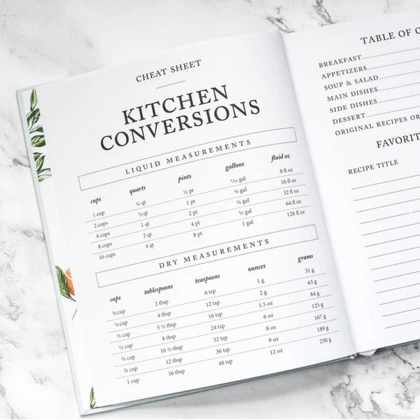An open recipe book showing printed kitchen conversion cheat sheets, including liquid and dry measurement tables, photographed on a light marble surface.