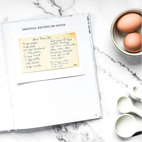 An open recipe book on a white marble surface, showing a handwritten recipe note on a yellowed paper page. A small bowl with two brown eggs and two white spoons sit to the right.