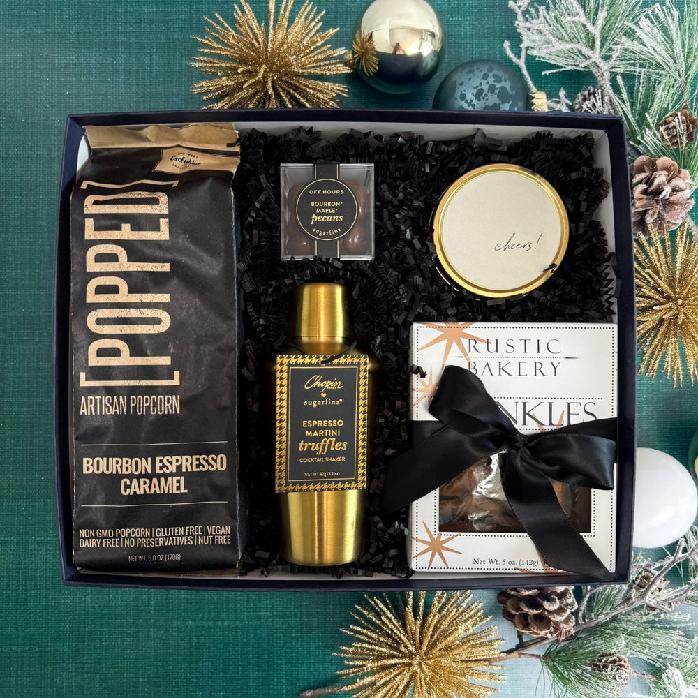 A curated gift box set on a green textured background with festive pine branches and gold star ornaments. The set includes a bag of Popped Artisan Popcorn, a gold shaker bottle of Sugarfina Martini Espresso truffles, a round gold Cheers tin candle, a box of Rustic Bakery Cinnamon Twinkles Cookies, and Sugarfina Bourbon Maple Pecans.