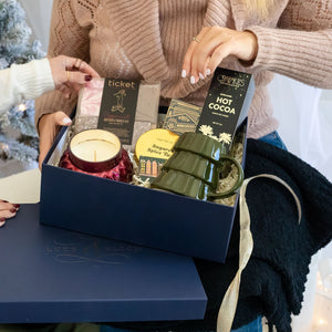 Woman opening a Fireside Christmas gift box with hot cocoa, mug, s'mores kit, decorative matchbook, playing cards, blanket and candle included, Christmas tree in the background