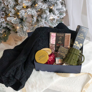 Fireside Luxury Curated Christmas Gift Box with candle, hot cocoa, mug, blanket, and more in front of a Christmas tree