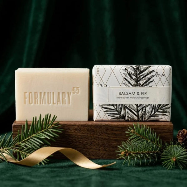 Two bars of Formulary 55 Balsam & Fir soap displayed on a wooden block against a dark green velvet background with pine branches and a gold ribbon. The left bar is a solid cream-colored soap embossed with 'FORMULARY 55'.