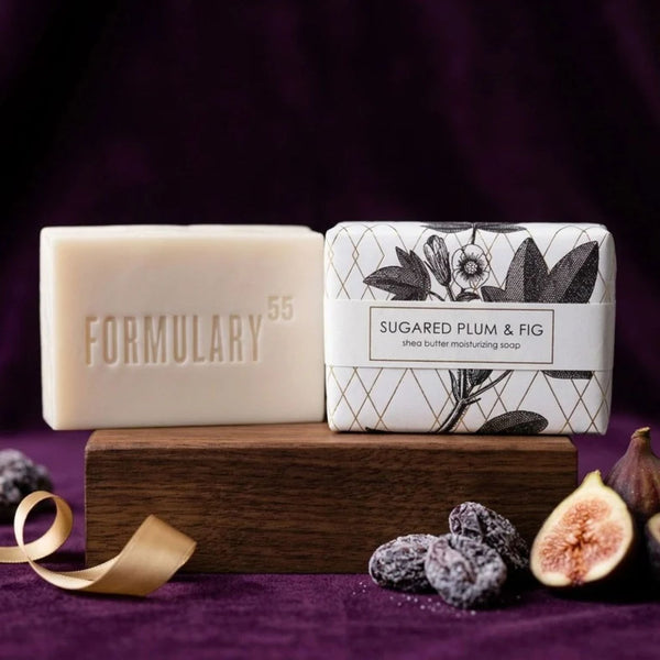 Two bars of FORMULARY 55 Sugared Plum & Fig Shea Butter Moisturizing Soap, one unwrapped in cream and one wrapped in white paper with black plum and fig illustrations, displayed on a wooden block with dried figs and a gold ribbon against a dark purple background.