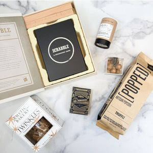A curated gift set arranged on a white marble surface, including a black Scrabble game box in an open beige gift box, a brown paper bag of Popped Artisan Truffle Popcorn, a Rustic Bakery Cinnamon Spice Twinkles Cookies, a bottle of bombay chai tea, and box of Monarch playing cards.