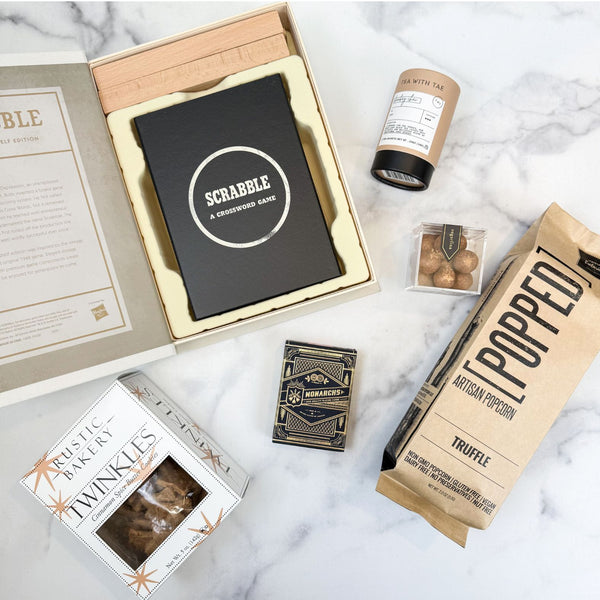 A curated gift set arranged on a white marble surface, including a black Scrabble game box in an open beige gift box, a brown paper bag of Popped Artisan Truffle Popcorn, a Rustic Bakery Cinnamon Spice Twinkles Cookies, a bottle of bombay chai tea, and box of Monarch playing cards.