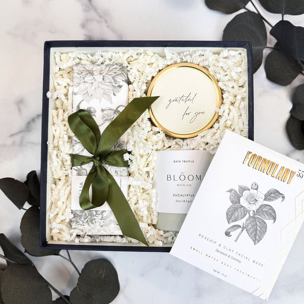 A curated gift box set on a white marble surface with dark green eucalyptus leaves. The set includes a square navy gift box filled with white crinkle paper, a gold-rimmed candle reading 'grateful for you', a white & sage green bath truffle labeled 'BLOOM BATH Eucalytus Bath Truffle', a white card labeled 'FORMULARY 55', and Lollia Elegance Shea Butter Handcreme with a dark green ribbon bow.
