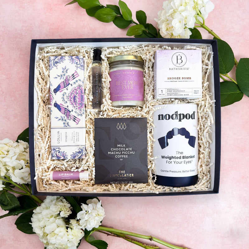 Joyful Retreat Luxury Curated Mother's Day Gift Box from Luxe & Bloom with various gift items including shea butter hand cream, lip balm, lavender oil roller, a candle, a chocolate bar, a snooze bath balm, and a Nodpod weighted eye mask.