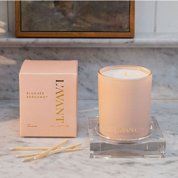 A Blushed Bergamot frosted glass jar candle with a single wick, featuring a soft blush pink color and gold foil branding that reads 'L'AVANT COLLECTIVE'. The candle is sitting next to it's box with matches laying next to it on a marble counter.