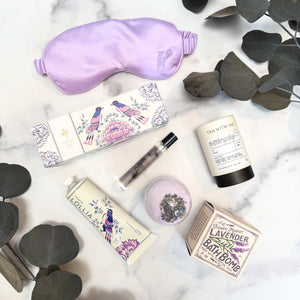A flat -lay of the items in our Lavender Blooms wellness gift box - featuring a lavender filled satin sleep mask, a lavender bath bomb, a small tranquil roller bottle, fruit & berry oolong tea, and a Lollia Imagine shea butter hand cream. The set is photographed from above on a white marble surface with dark eucalyptus leaves around it.