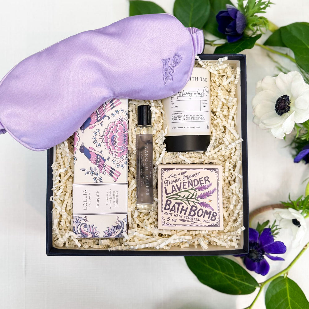 Lavender Blooms wellness gift box arranged in a navy gift box filled with ivory shredded paper, featuring a lavender filled satin sleep mask, a lavender bath bomb, a small tranquil roller bottle, and a Lollia Imagine shea butter hand cream. The set is photographed from above on a white linen surface with green leaves and flowers around it.