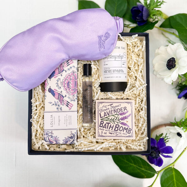 Lavender Blooms wellness gift box arranged in a navy gift box filled with ivory shredded paper, featuring a lavender filled satin sleep mask, a lavender bath bomb, a small tranquil roller bottle, and a Lollia Imagine shea butter hand cream. The set is photographed from above on a white linen surface with green leaves and flowers around it.