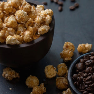 Popped Bourbon Espresso Caramel Artisan Popcorn in a dark bowl with scattered caramel popcorn pieces and a small bowl of coffee beans on a dark surface.