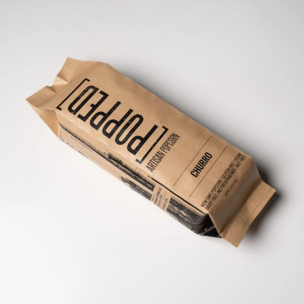 A kraft paper bag of Popped Artisan Popcorn in Churro flavor, photographed at an angle on a clean white background. Build a luxury custom gift box with Luxe & Bloom.
