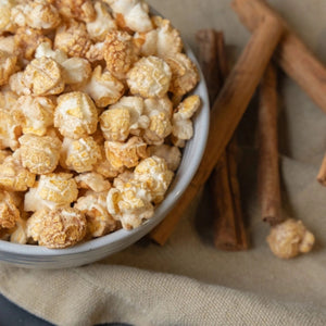Popped churro artisan popcorn in a white bowl with cinnamon sticks on a beige fabric placemat.