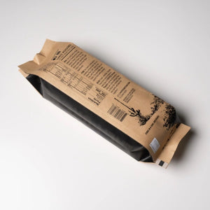 A kraft paper bag package of Popped  Churro Artisan Popcorn, photographed on a clean light gray background. The package is shown at an angle, displaying the printed back panel with text, a barcode, and a small cactus illustration.