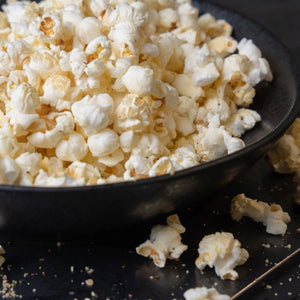 A close-up of fluffy Popped Truffle Artisan Popcorn piled in a black bowl on a dark surface, with a few pieces scattered around.