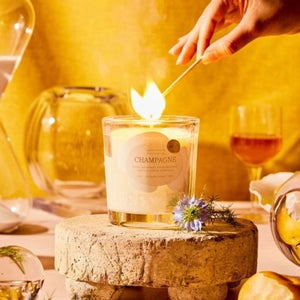 Rewined Champagne Candle 6 oz candle in a clear glass jar with gold writing and accents being lit with a match on a table setting with glasses and flowers.