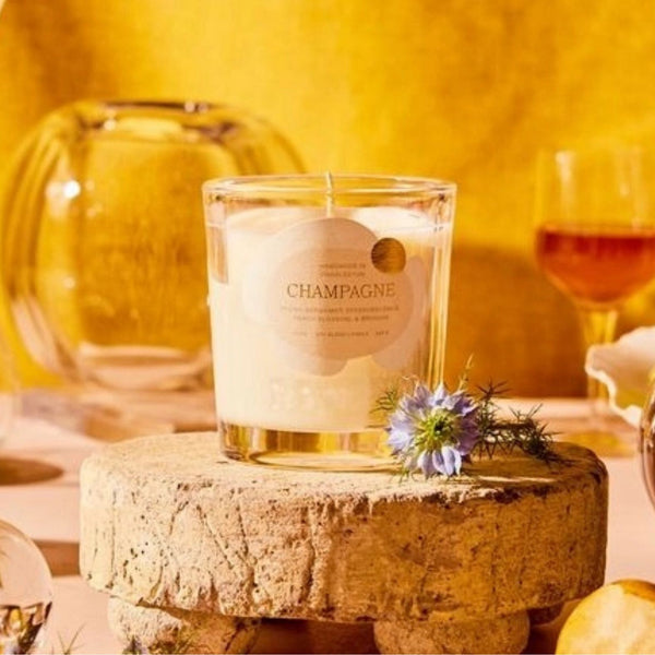 A champagne-scented candle in a clear glass jar sits on a rustic wooden block, styled with a small purple flower. The background is a warm, golden-yellow tone with a glass bowl and a wine glass in soft focus.