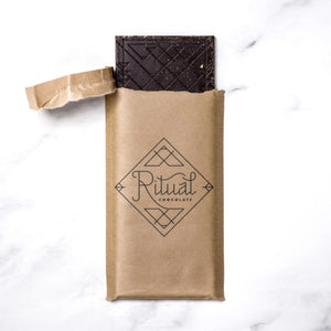 A partially unwrapped dark chocolate bar in a brown kraft paper sleeve with the brand name 'Ritual Chocolate' printed on the front, set on a white marble background.