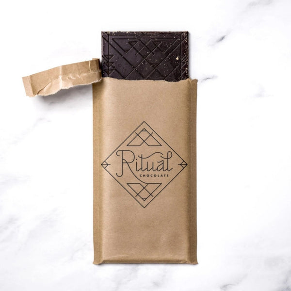A partially unwrapped dark chocolate bar in a brown kraft paper sleeve with the brand name 'Ritual Chocolate' printed on the front, set on a white marble background.