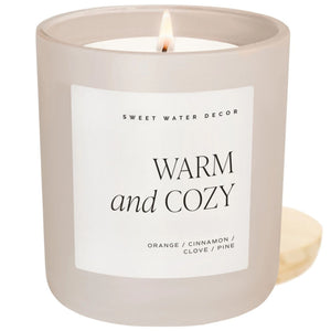 A frosted glass jar candle with a lit wick, featuring a white label that reads 'SWEET WATER DECOR WARM and COZY' and 'ORANGE / CINNAMON / CLOVE / PINE'. A wooden lid is visible to the right of the candle on a white background.