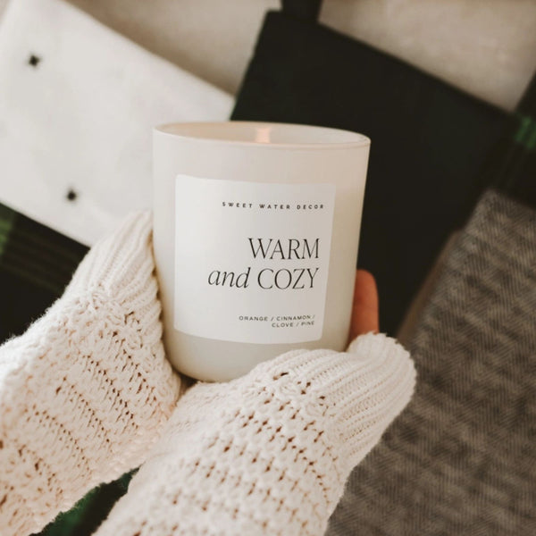 Hands with cozy cream sweater sleeves holding Sweet Water Decor Warm & Cozy 15 oz Matte Jar Candle with a cozy background of textured fabrics | Build A Luxury Custom Gift Box with Luxe & Bloom