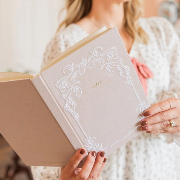 Woman holding a Selah Publications Lainey notebook with intricate white stitched design on linen colored cover  on a blurred background