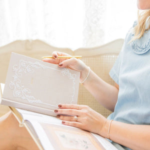Woman sitting on a couch holding an open Selah Publications Lainey notebook with a linen cover with white stitching details.