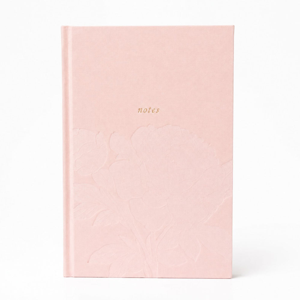 A woman holding The Poly Blair Notebook with a blush pink hardcover with the word 'notes' printed in gold on the cover, photographed in a softly lit indoor setting.