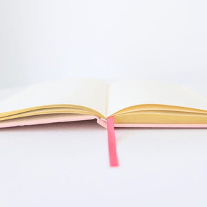 An open notebook with cream-colored pages, yellow-gold inner edges, a pale pink cover, and a bright pink ribbon bookmark, set against a clean white background.